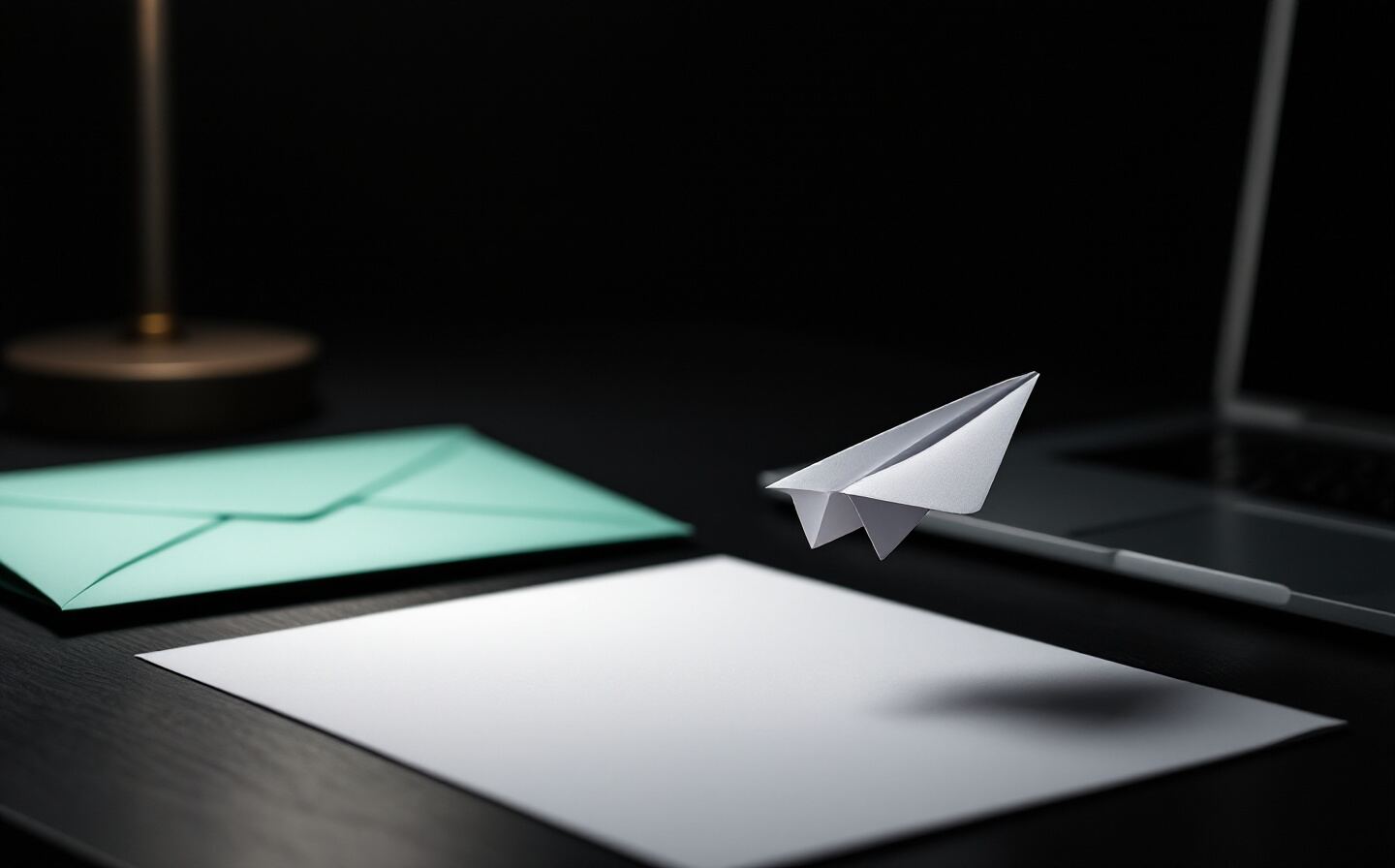 Un avion en papier blanc s'envole au-dessus d'un bureau sombre, à côté d'une enveloppe vert menthe et d'un ordinateur portable.