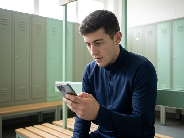 Tom, a young club player in a navy jacket, looks at a notification on his phone in the changing room.